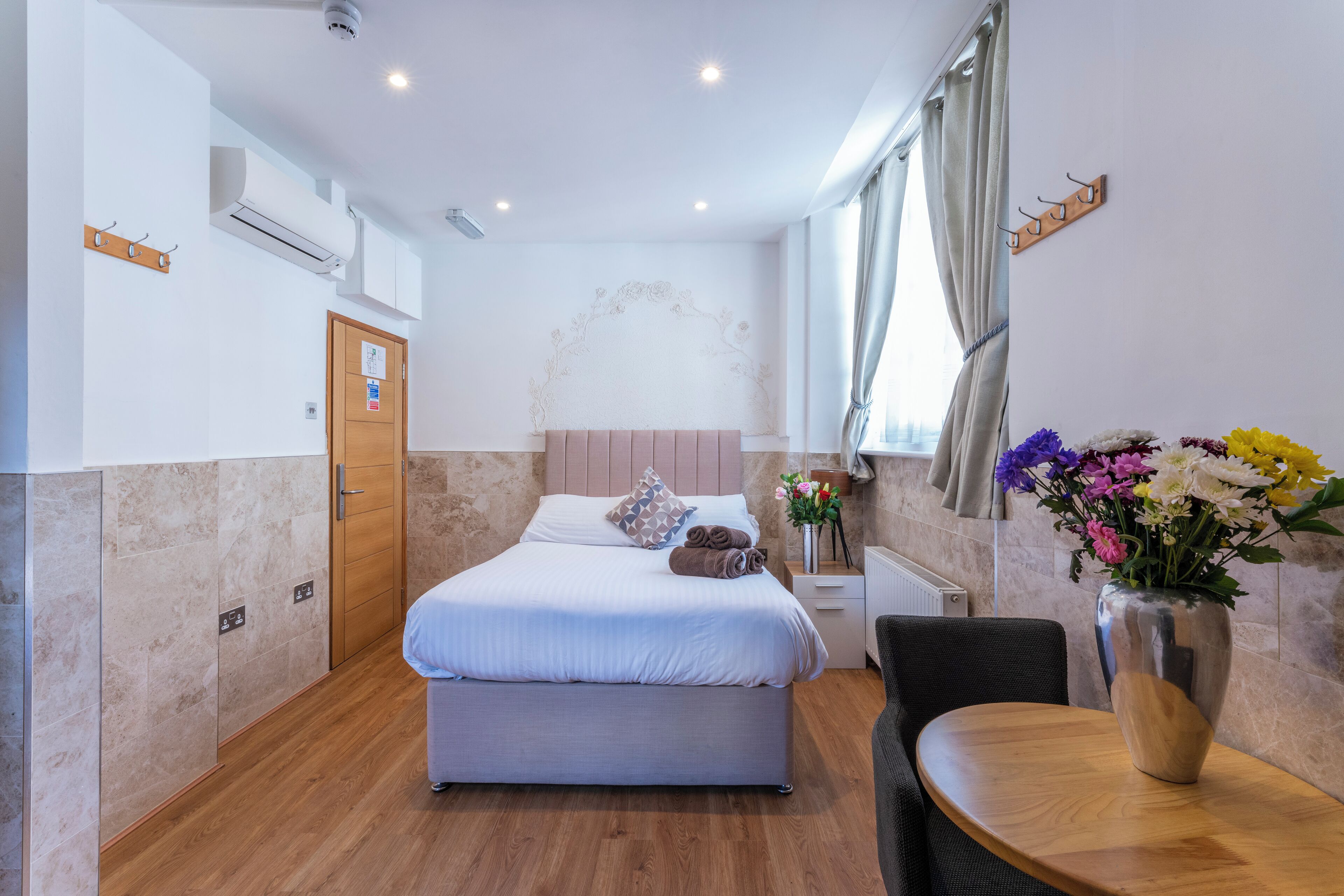 A modern hotel room with a bed, a wooden table, a chair, a vase with flowers, and a window with curtains.