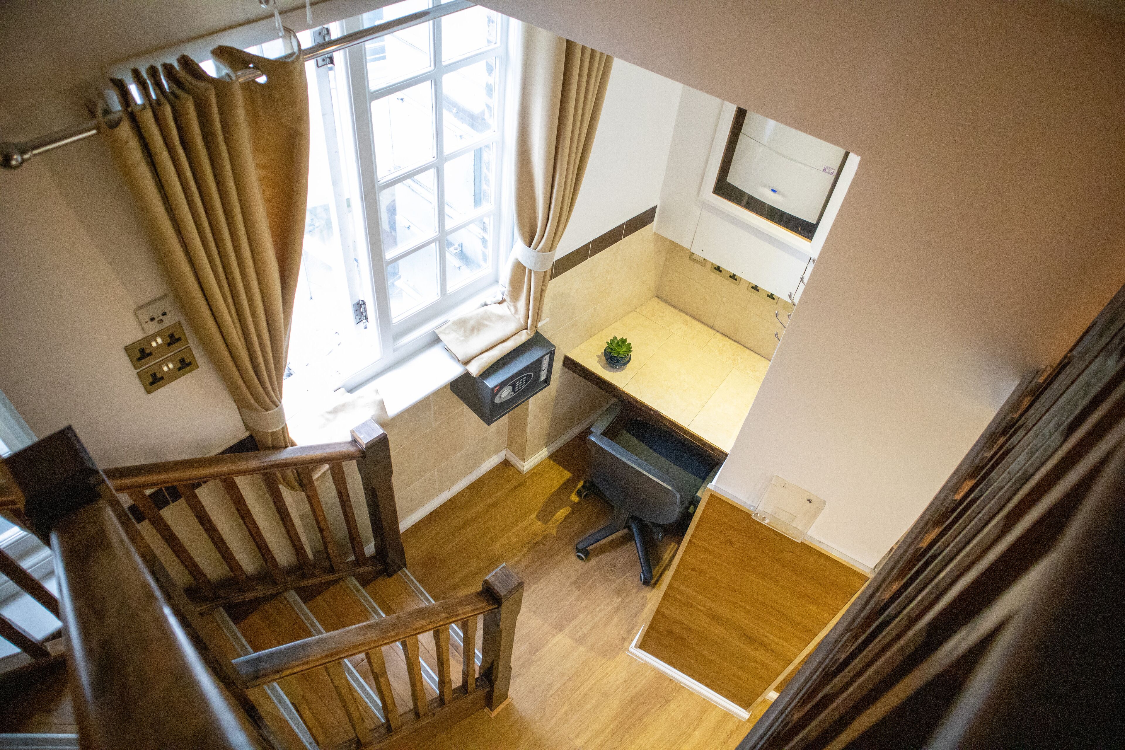 Een kleine kamer met een houten trap, een bureau met een computer, een klein plantje en een raam met gordijnen.