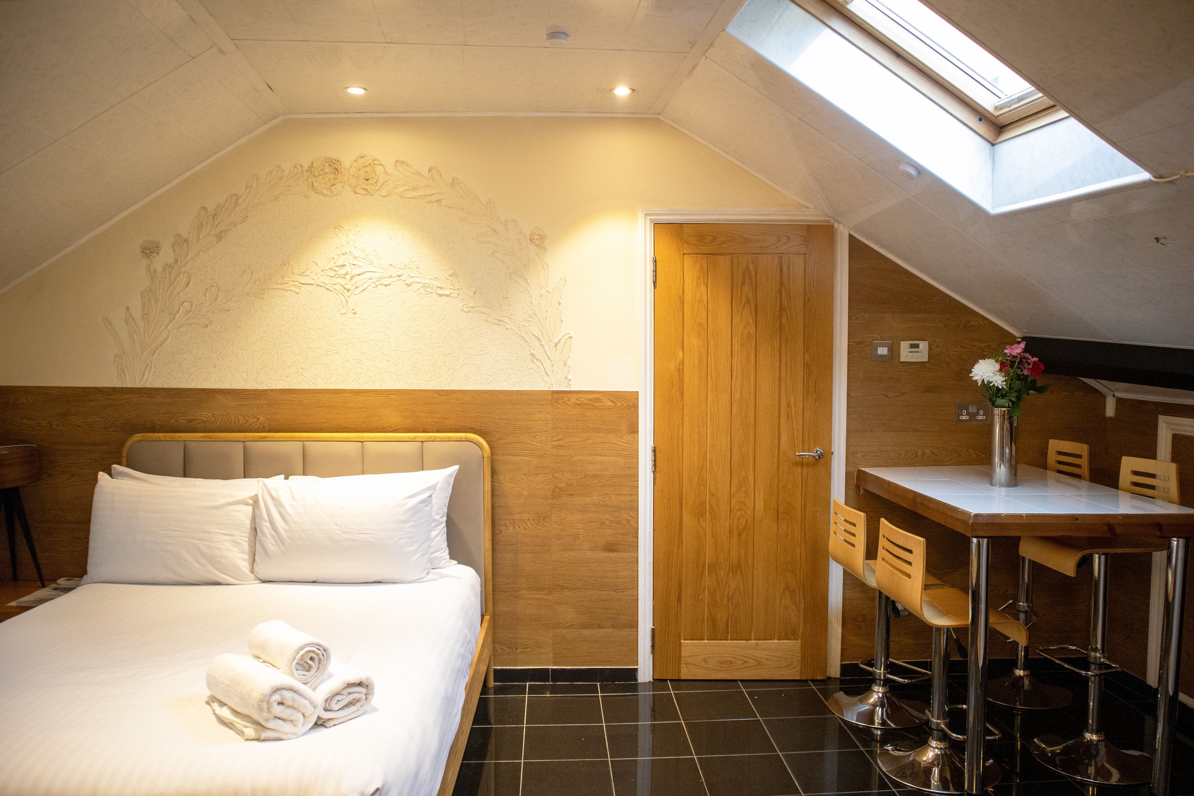 A hotel room with a bed, a desk with chairs, a skylight, and a wooden door.