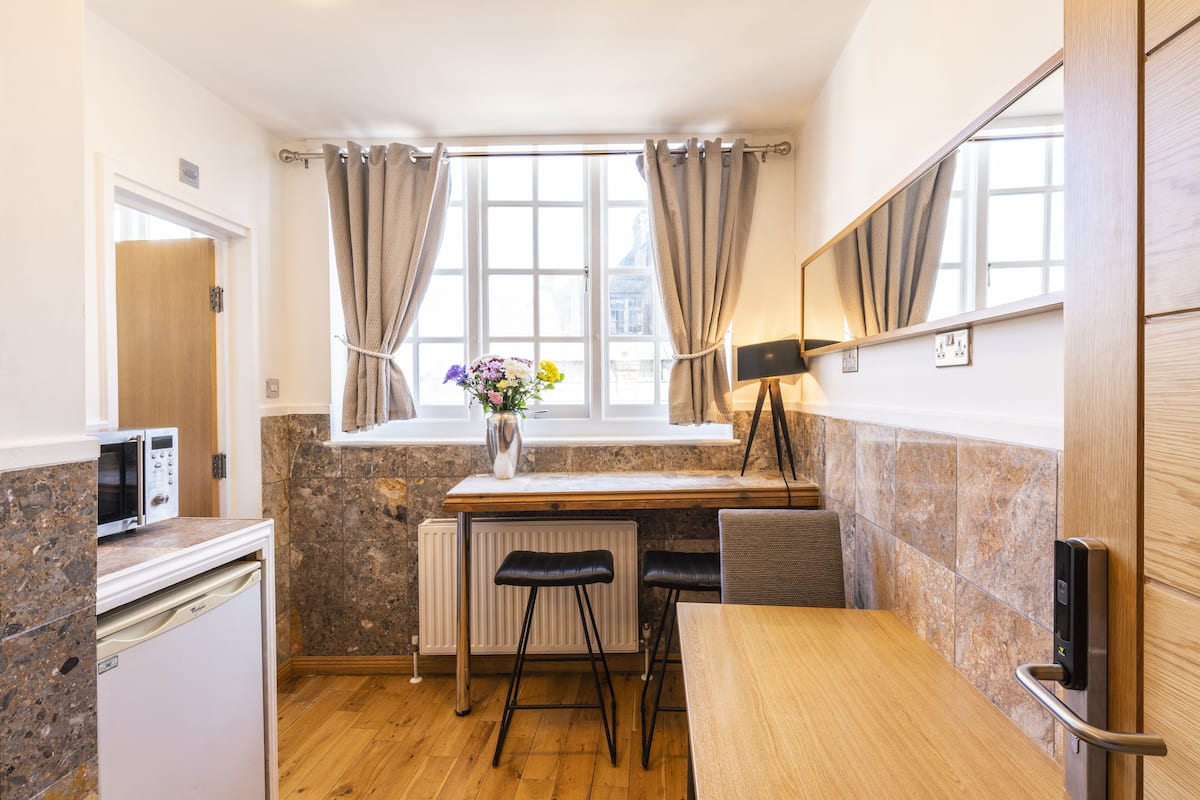 A compact kitchen with a microwave, a small refrigerator, a wooden table, two bar stools, a radiator, and a window with curtains.