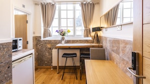 Une cuisine compacte équipée d’un four à micro-ondes, d’un petit réfrigérateur, d’une table en bois, de deux tabourets de bar, d’un radiateur et d’une fenêtre avec des rideaux.