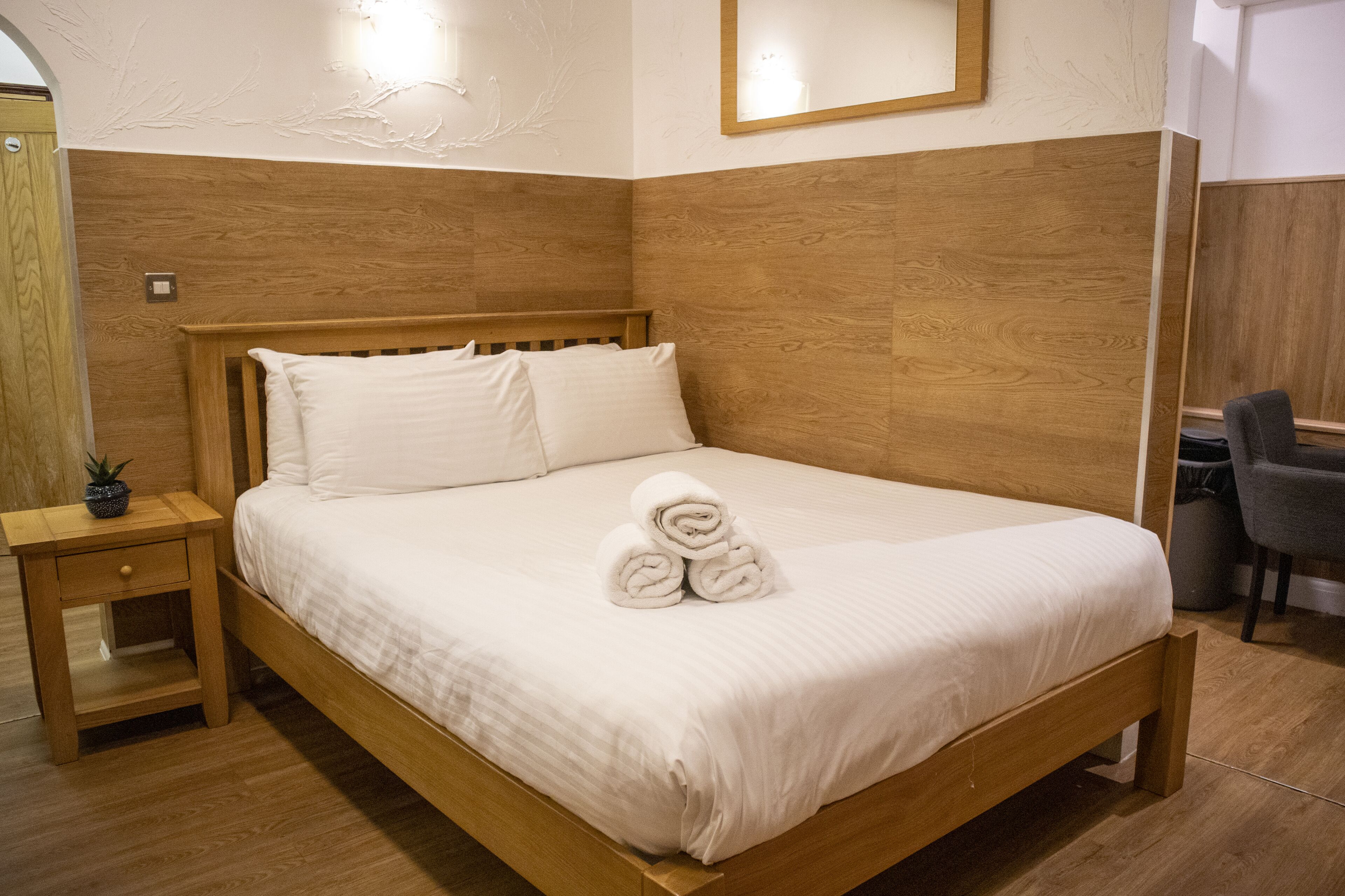 A neatly made bed with white linens and rolled towels, a wooden nightstand with a drawer, and a small potted plant.
