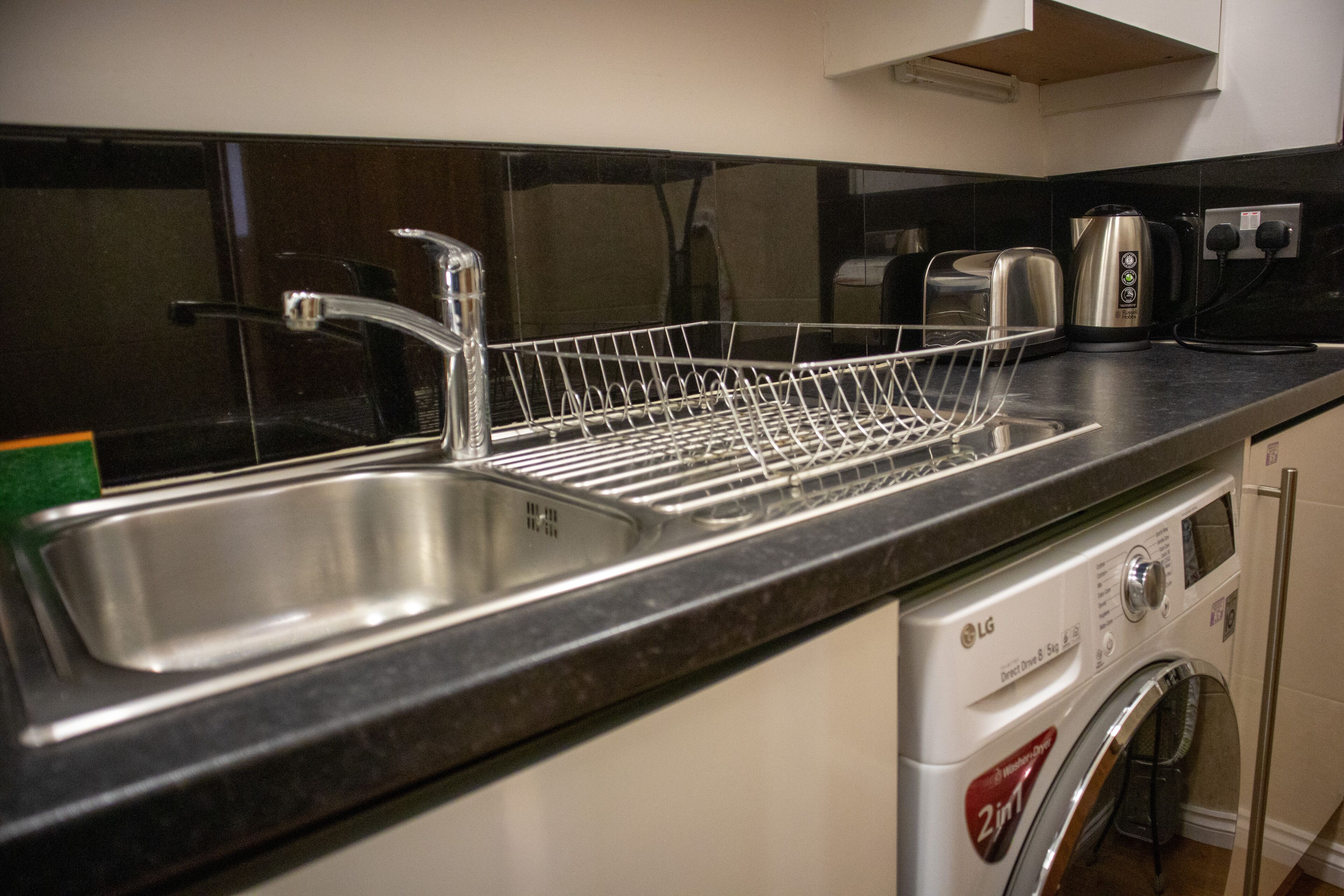 A kitchen with a stainless steel sink, a dish rack, a toaster, and a washing machine.