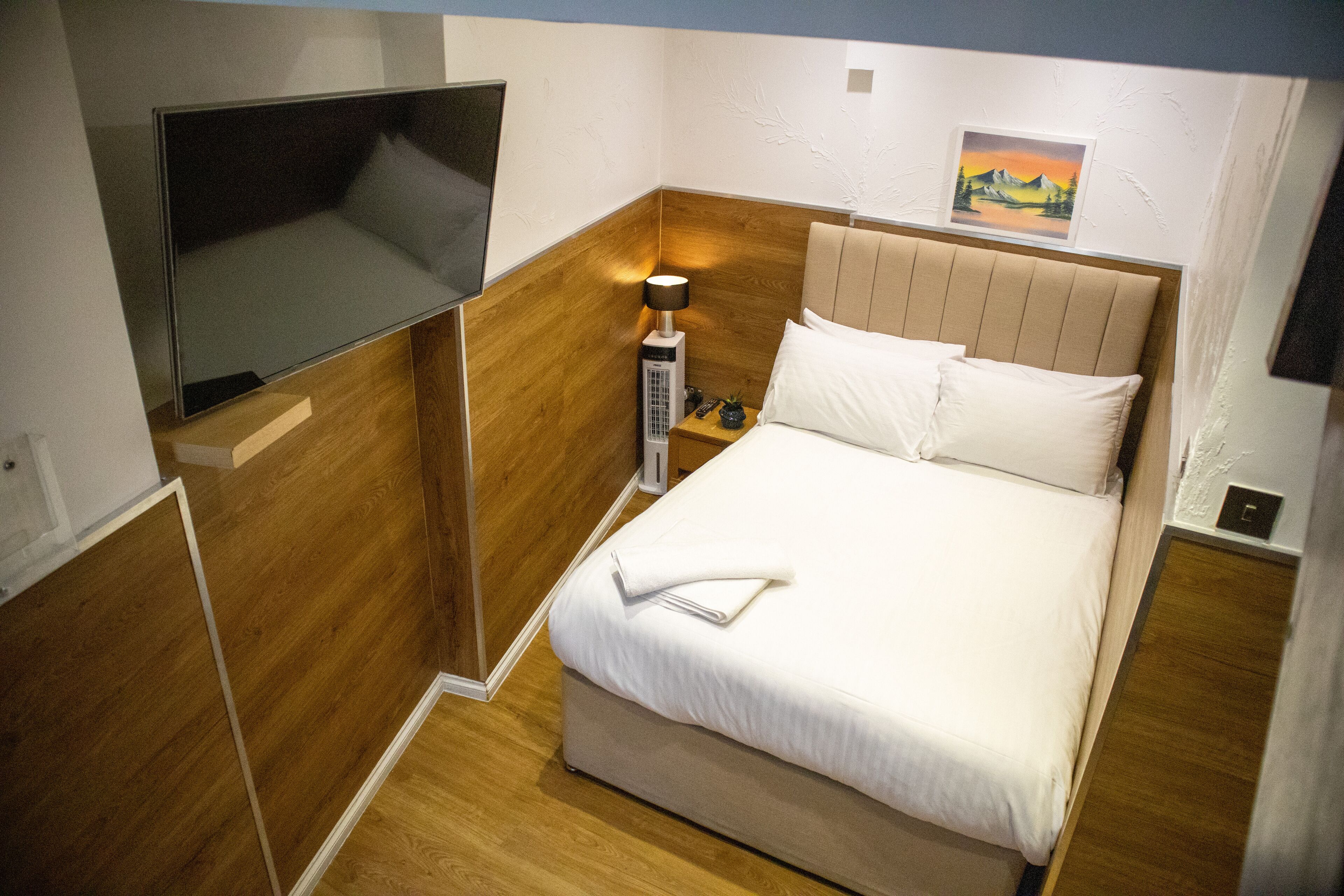 A compact hotel room with a bed, a bedside table, a lamp, and a wall-mounted TV.