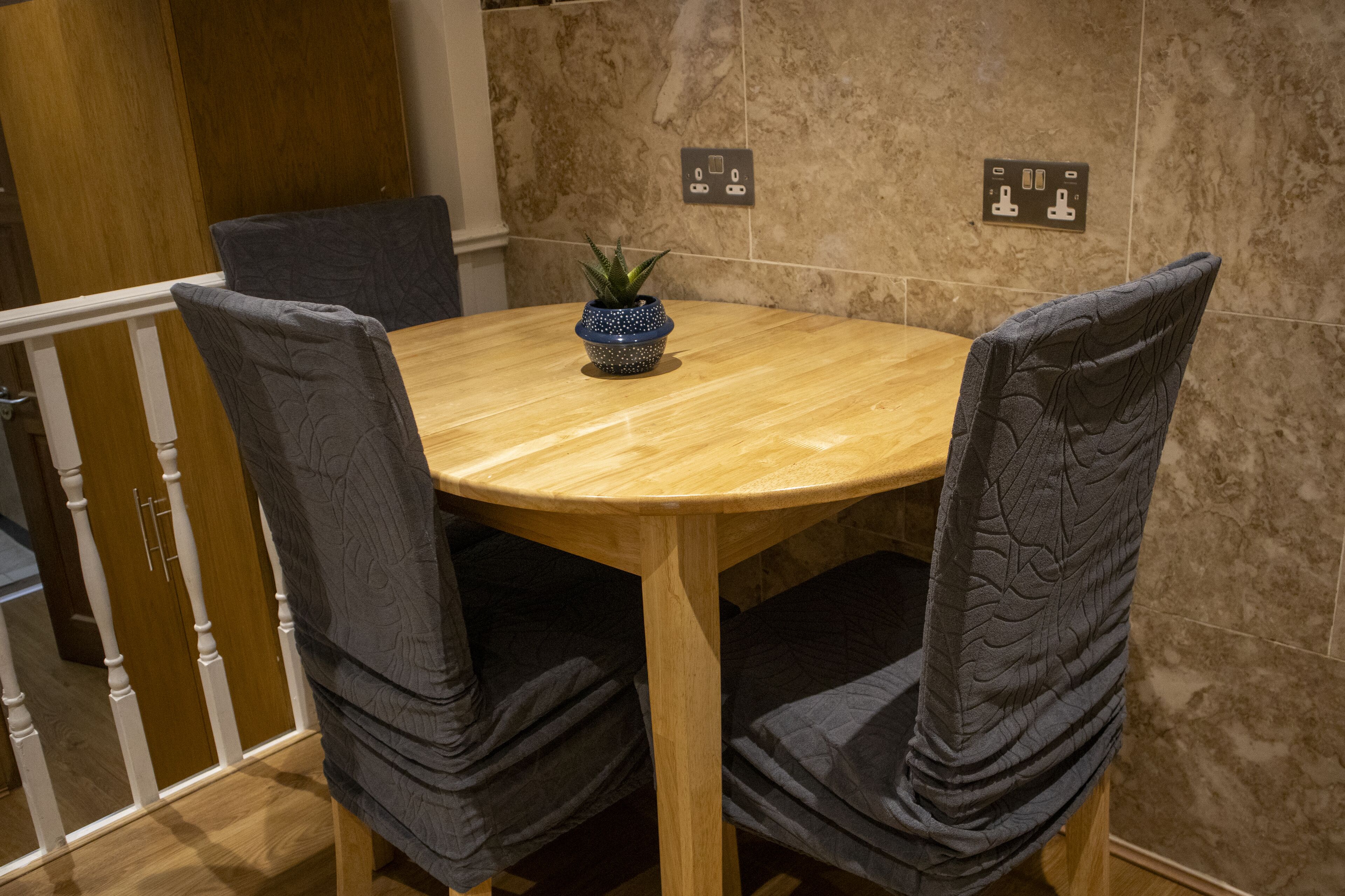 A round wooden table with four upholstered chairs, a small potted plant, and electrical outlets on the wall.