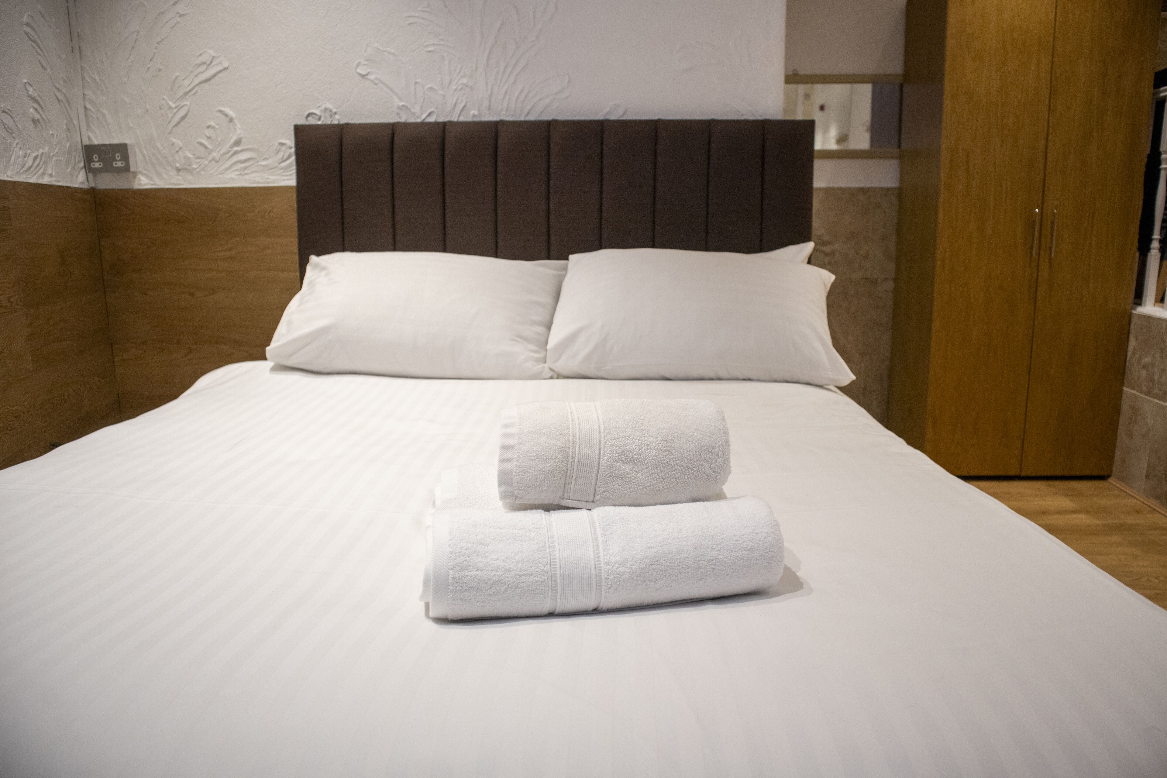 Una cama bien hecha con sábanas blancas y dos toallas enrolladas, un cabecero de madera y un armario de madera en una habitación de hotel.