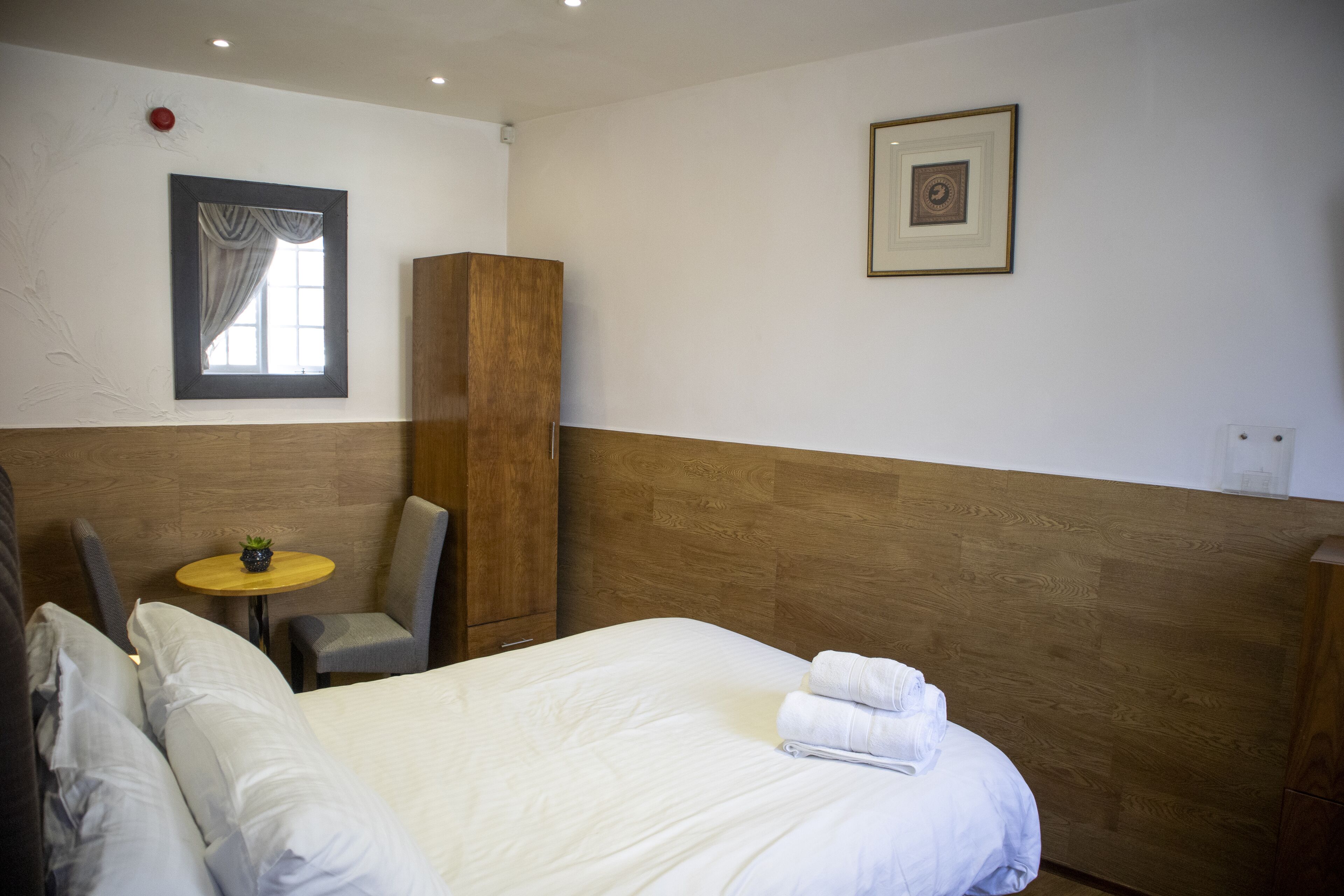 Habitación de hotel con cama, una silla, una mesita con una planta y un cuadro en la pared.