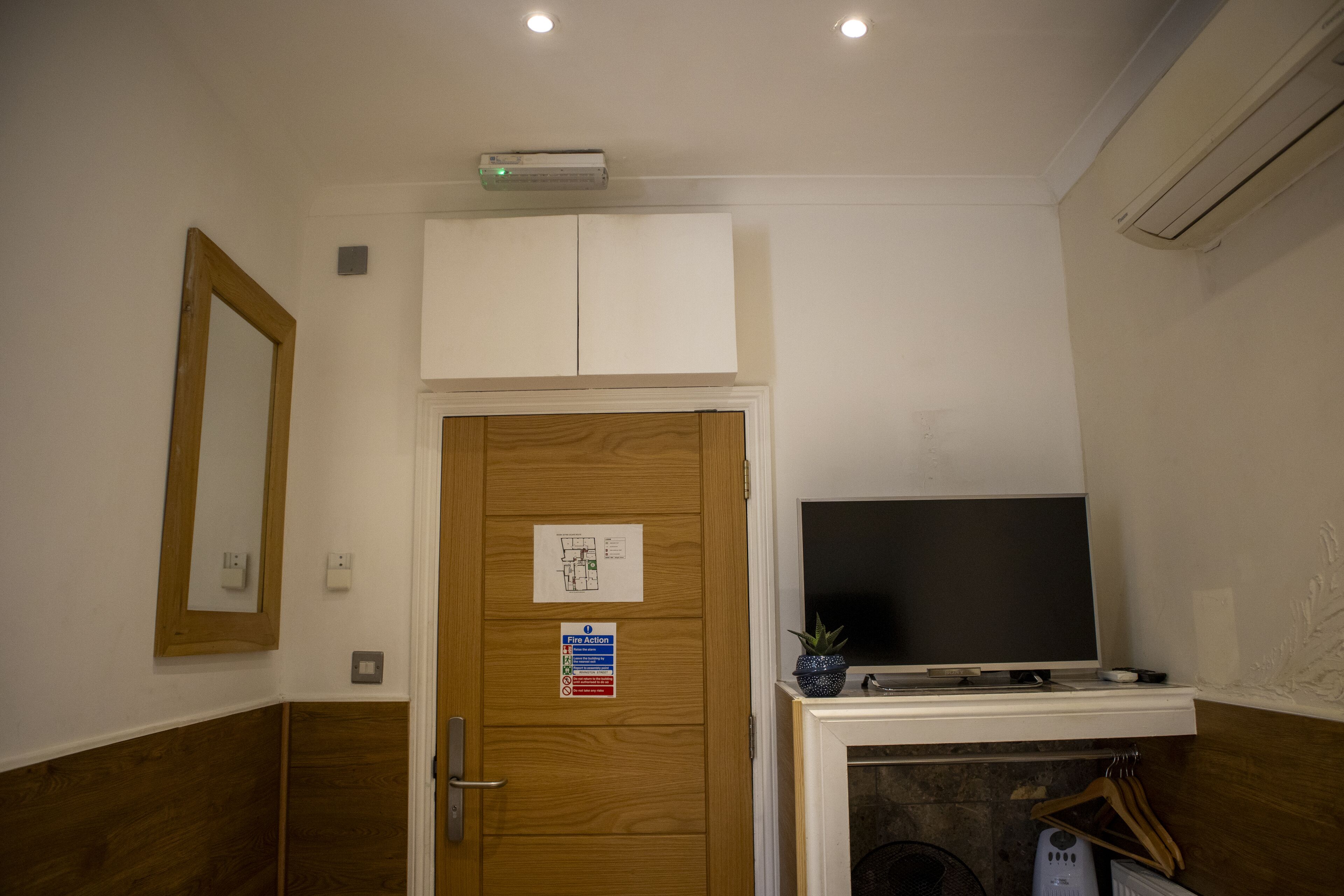 A hotel room with a wooden door, a fire action sign, a flat-screen TV, and a small plant on the mantel.