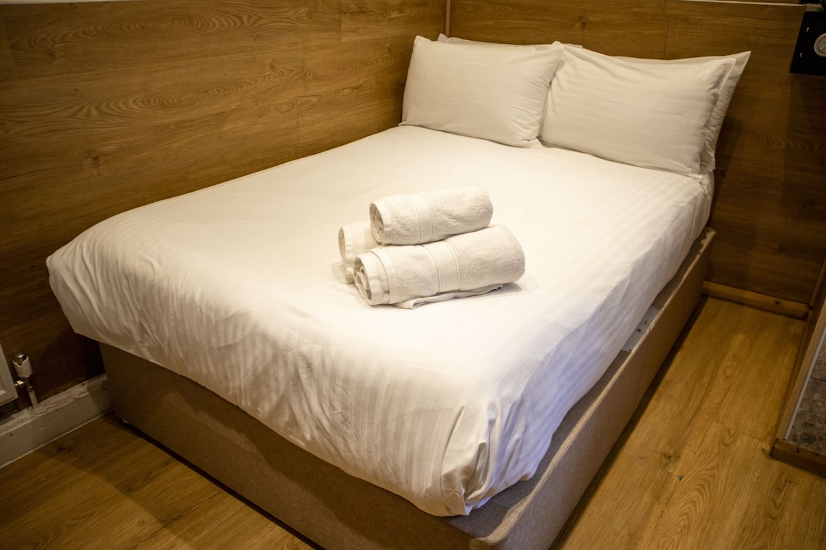 A neatly made bed with white linens and two rolled towels on top, set in a room with wooden flooring and headboard.