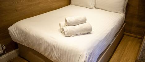 A neatly made bed with white linens and two rolled towels on top, set in a room with wooden flooring and headboard.