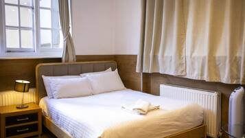A neatly made bed with white linens, a bedside lamp, a wooden nightstand, and a window with curtains.