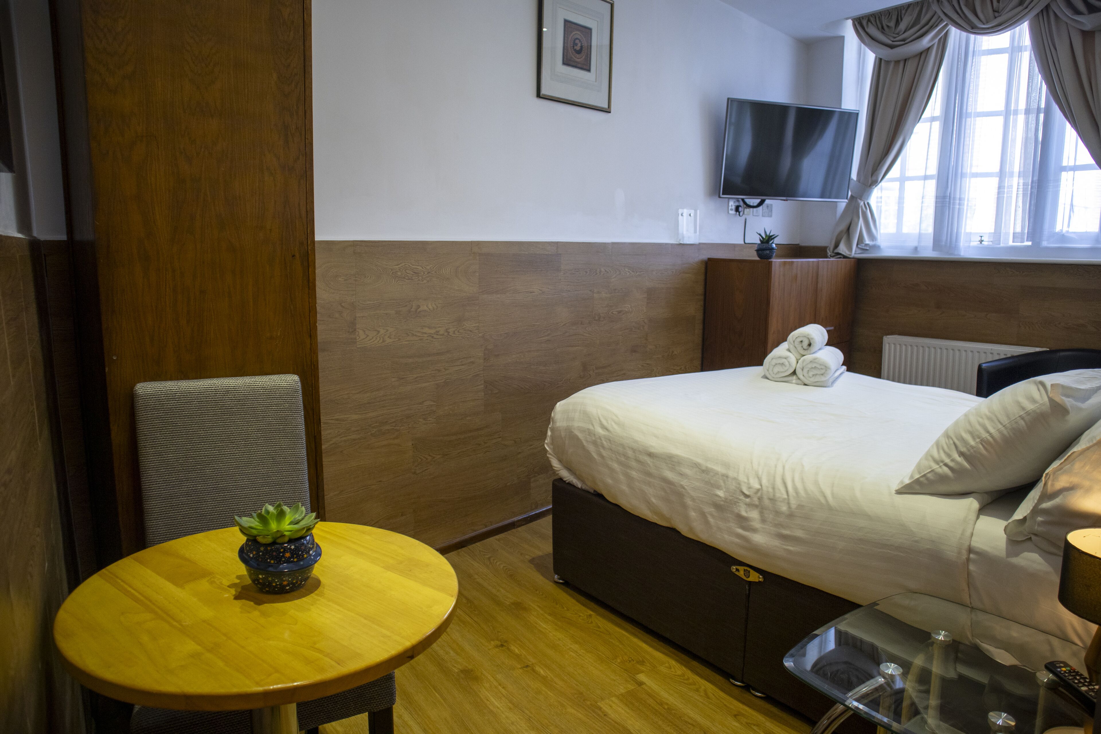 Een hotelkamer met een bed, een televisie, een klein tafeltje met een plant en een raam met gordijnen.