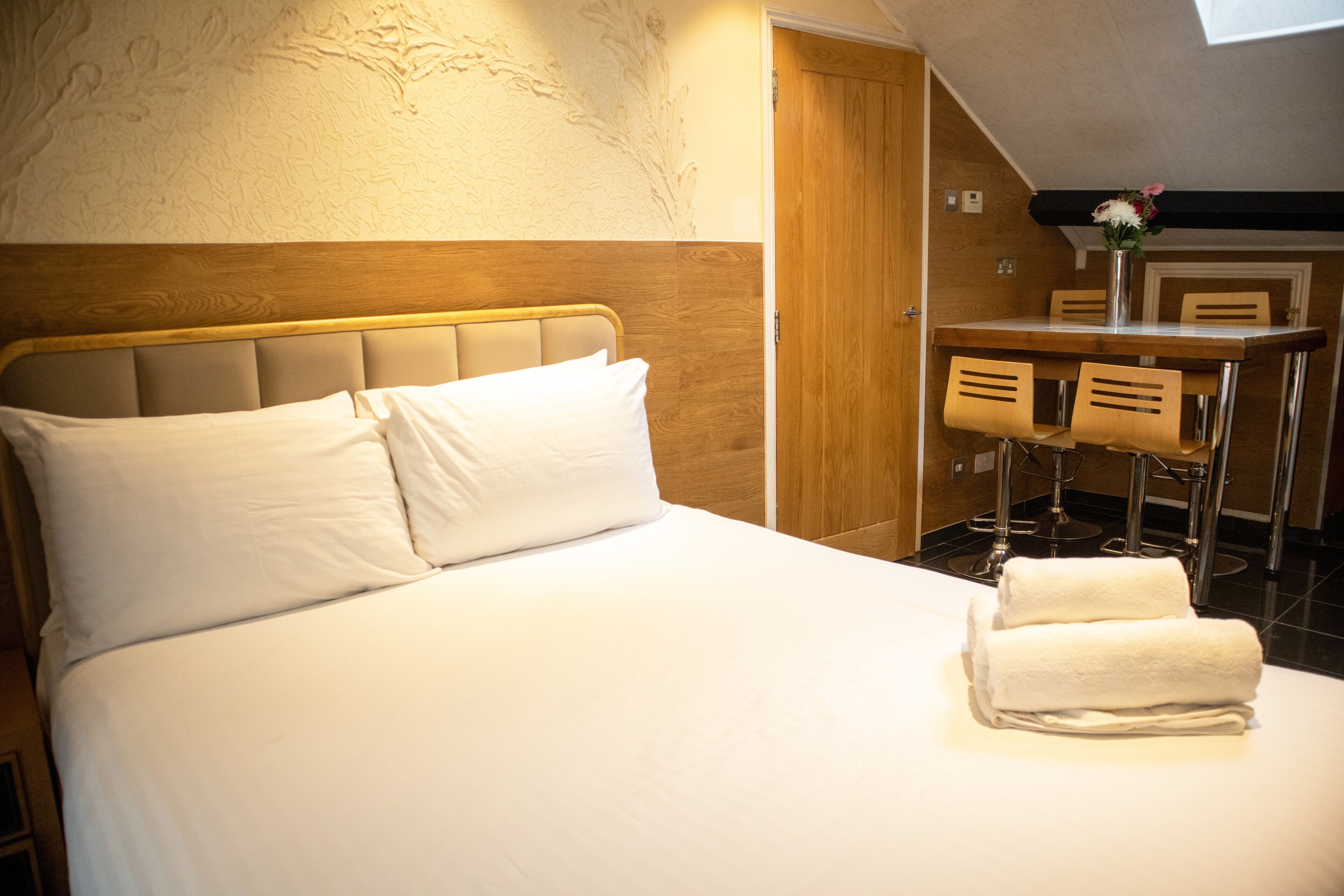 A neatly made bed with white linens and pillows, a wooden headboard, and a small table with chairs in the background.