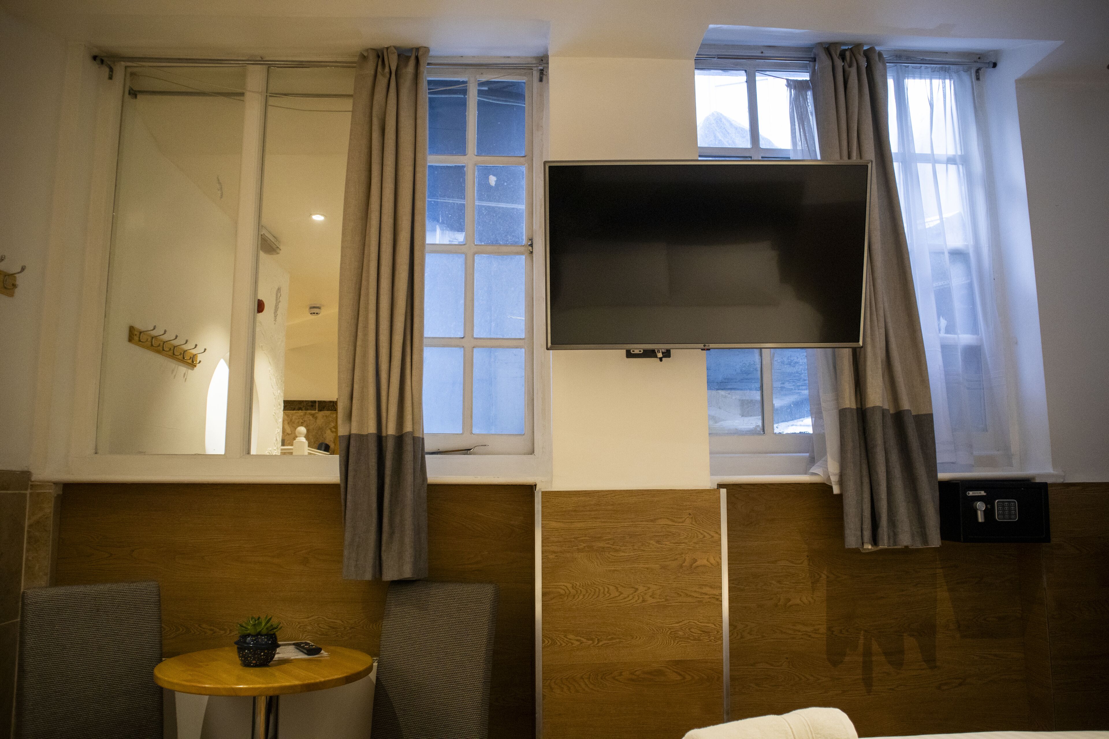 Een kamer met een groot raam, een flatscreen televisie aan de muur, een klein rond tafeltje met een plant en een bank.