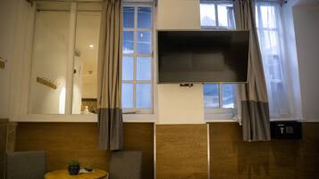 Een kamer met een groot raam, een flatscreen televisie aan de muur, een klein rond tafeltje met een plant en een bank.