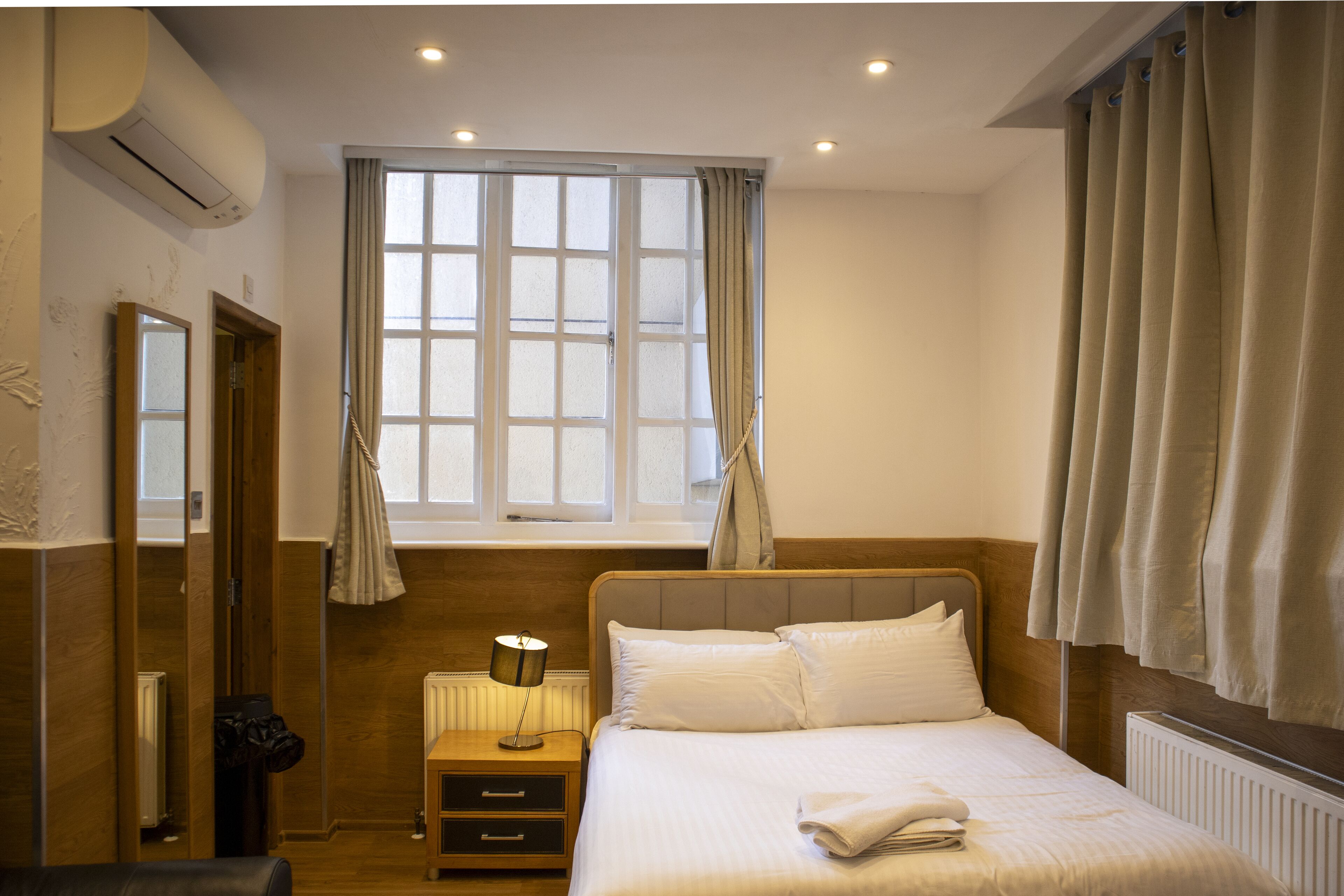 A bedroom with a bed, a nightstand, a lamp, a window with curtains, and a radiator.
