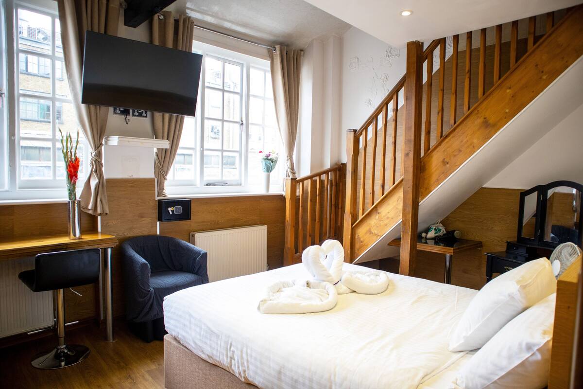 A hotel room with a bed, a desk with a chair, a TV mounted on the wall, a wooden staircase, and a window with curtains.