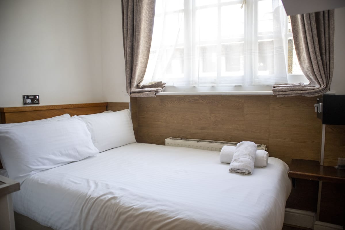 A hotel room with a wooden headboard, a bed with white linens, a window with curtains, and a bedside table with a lamp.