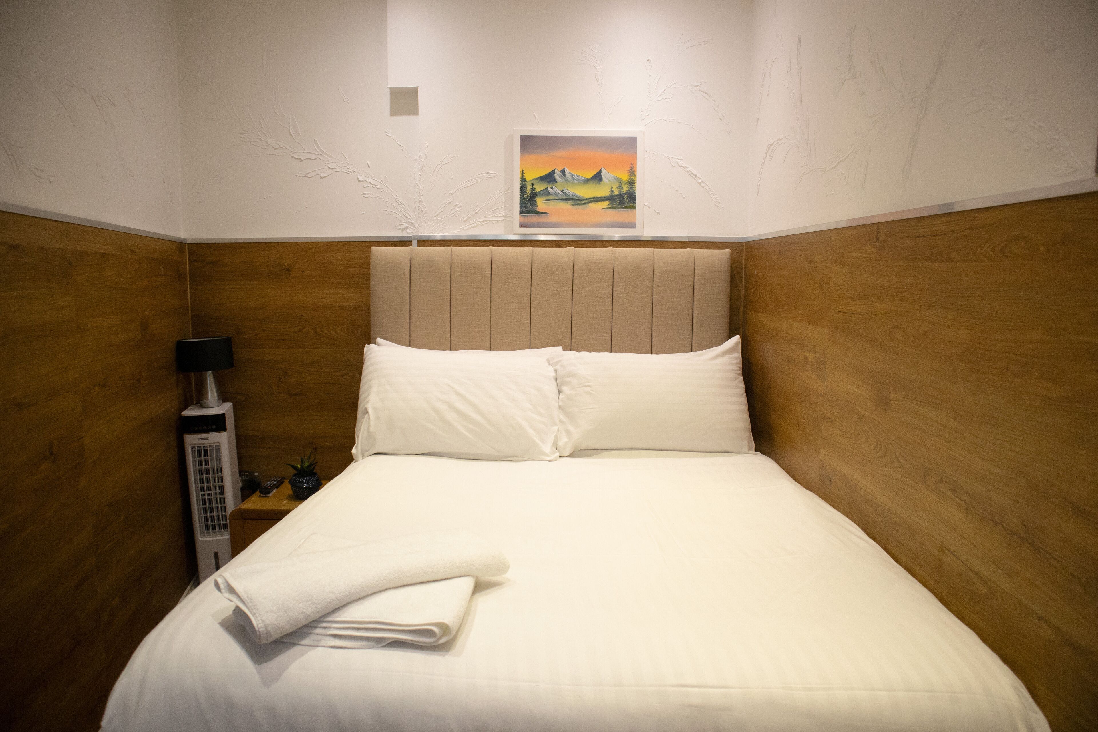 Una cama bien hecha con ropa blanca y una toalla doblada, una mesita de noche con una lámpara, y un cuadro de un paisaje de montaña en la pared.