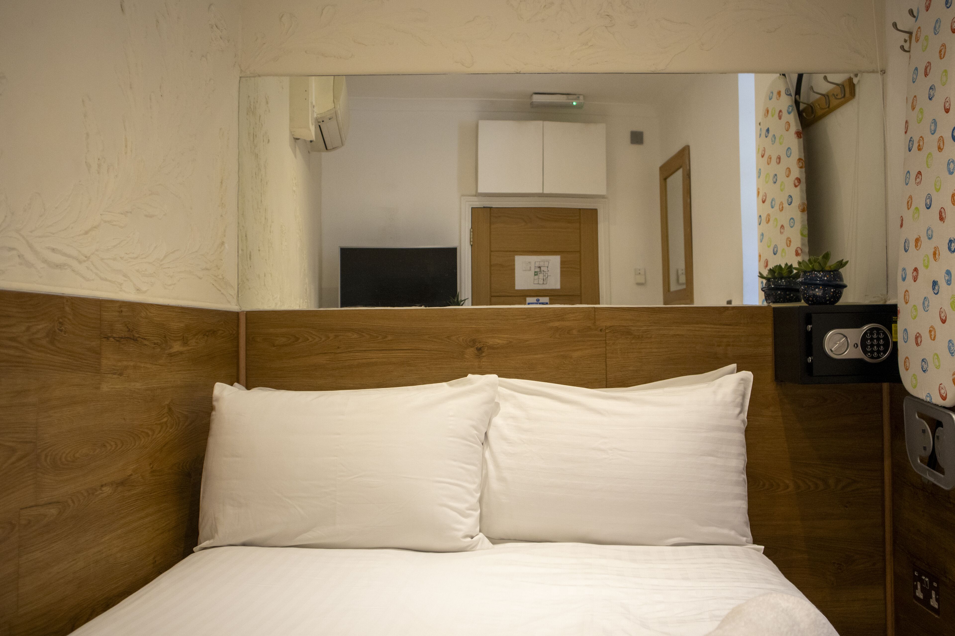 Quarto de hotel com cabeceira de madeira, cama com roupa de cama branca, uma pequena mesa com uma planta em vaso e um espelho refletindo o quarto.