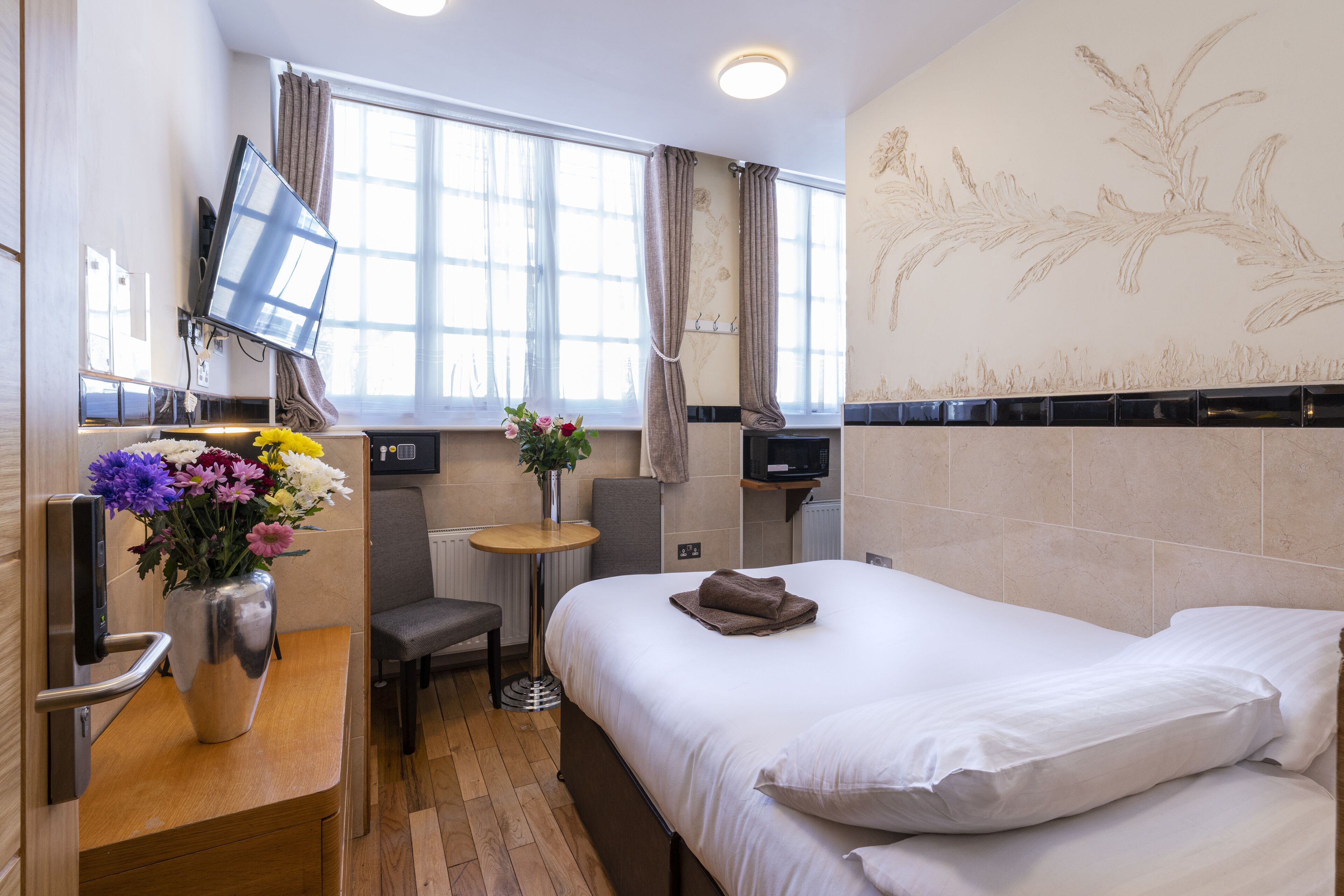 A hotel room with a bed, a desk with a vase of flowers, a chair, a TV mounted on the wall, and a window with curtains.