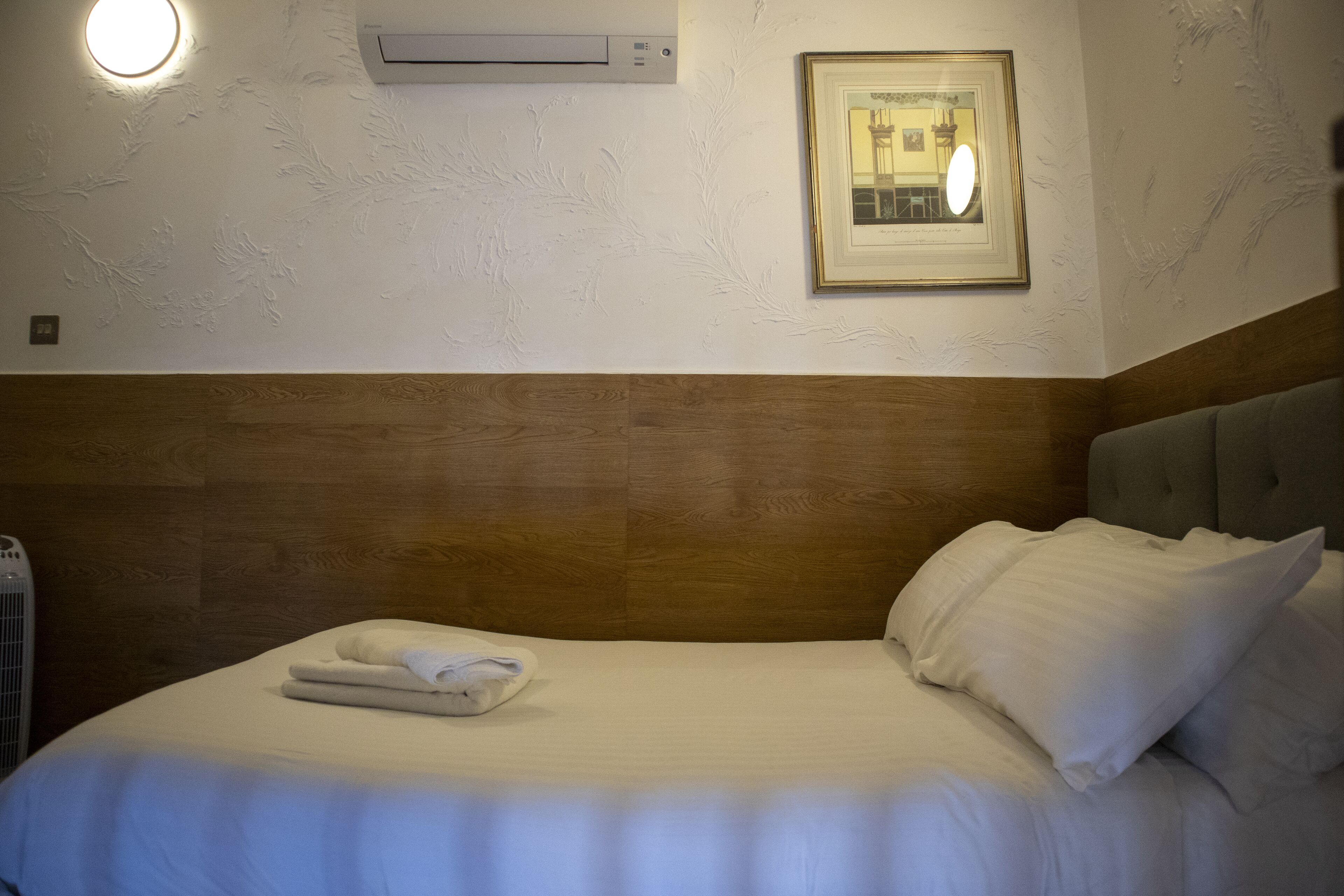 A hotel room with a wooden headboard, a neatly made bed with white linens, a framed picture on the wall, and an air conditioning unit.