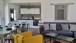 Een moderne woonkamer met een grijze bank, een gele fauteuil en een glazen salontafel. Op de achtergrond is een eethoek met een glazen tafel en stoelen zichtbaar.