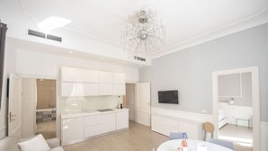 A modern living room with a white sofa, a dining table with a bottle and glass, and a kitchen area with white cabinets and a chandelier. - St. Peter Beautiful 2BR Apartment (Roma)