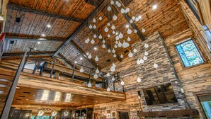 A spacious interior with wooden beams and stone accents, featuring a high ceiling with hanging light fixtures and a large window providing natural light. - Fox Trot Lodge - 6 King Suites - Pickleball Court (Broken Bow)