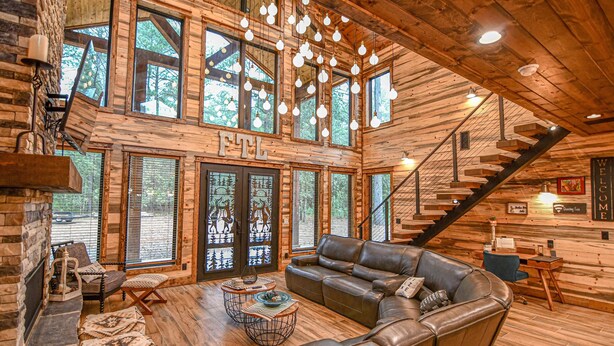 A spacious log cabin interior with a large leather sofa, a fireplace, and a staircase leading to an upper level. - Fox Trot Lodge - 6 King Suites - Pickleball Court (Broken Bow)