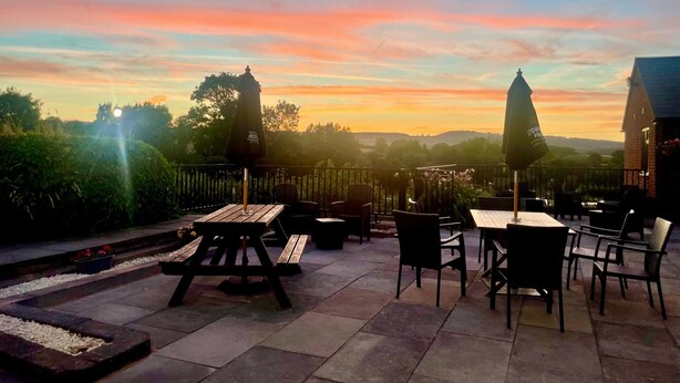 An outdoor patio area with wooden tables and chairs, umbrellas, and a view of the sunset over a hilly landscape.