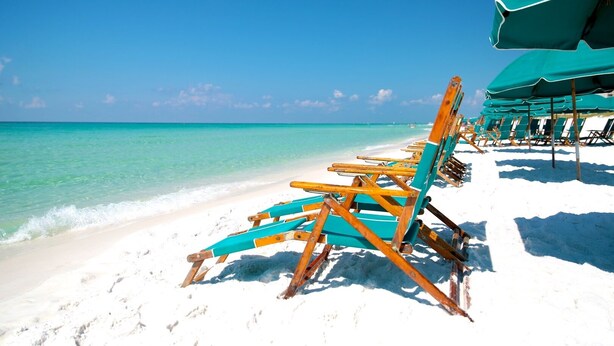 Ein Strand mit Reihen aus Holzliegen und Sonnenschirmen, bewölkter Himmel und türkisblauem Wasser.