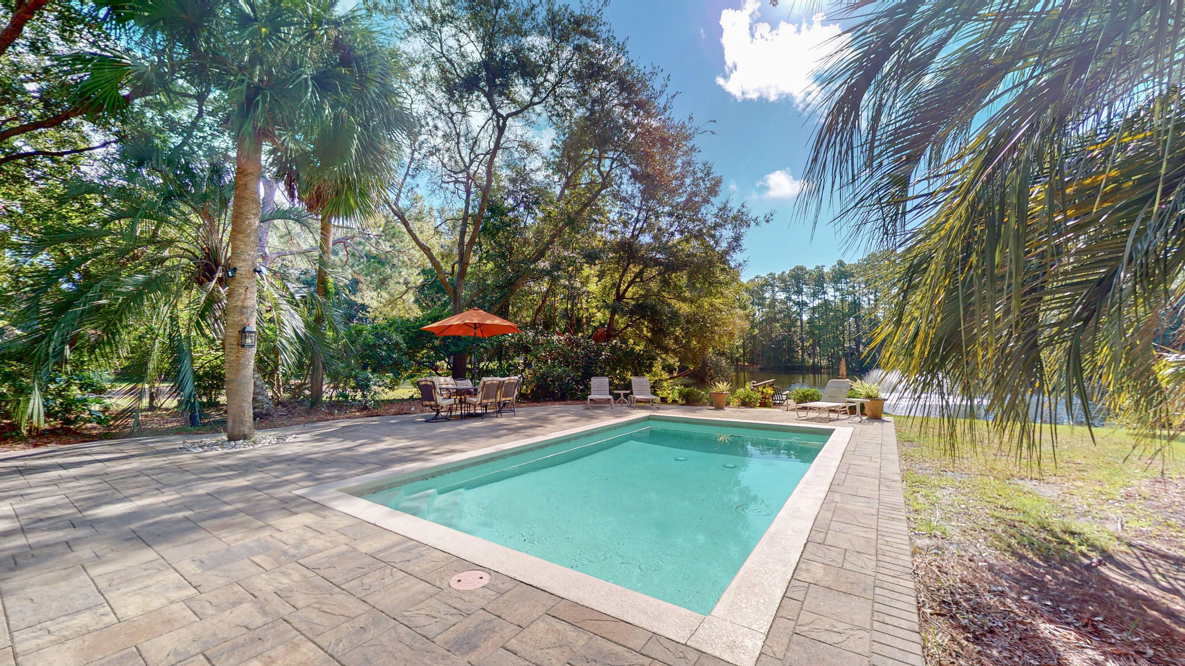 A rectangular swimming pool with a tiled deck, surrounded by lush greenery and trees.