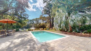 A rectangular swimming pool with a tiled deck, surrounded by lush greenery and trees.
