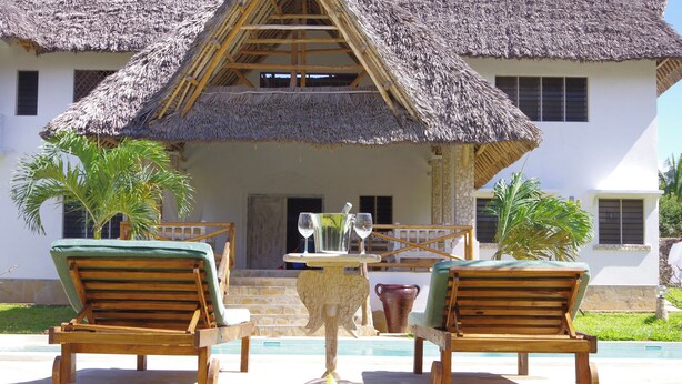 A thatched-roof house with a pool, two wooden lounge chairs, and a table with glasses.