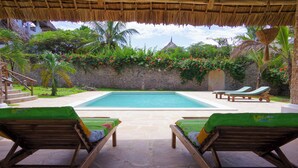 A pool area with two wooden lounge chairs, a stone pathway, and a thatched roof structure.