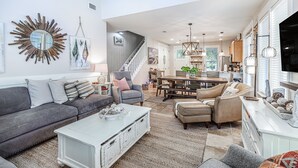 A spacious living room with a gray sofa, a white coffee table, and a dining area with a wooden table and chairs. A staircase leads to the upper floor, and a large mirror hangs on the wall. - GolfCourseView~6 Person GolfCart~HeatedPool*~Hot tub~FREE GOLF+Pet considered (Miramar Beach)
