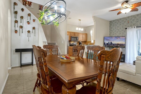 Uma sala de jantar com mesa e cadeiras de madeira, um candelabro e uma televisão exibindo uma vista urbana.