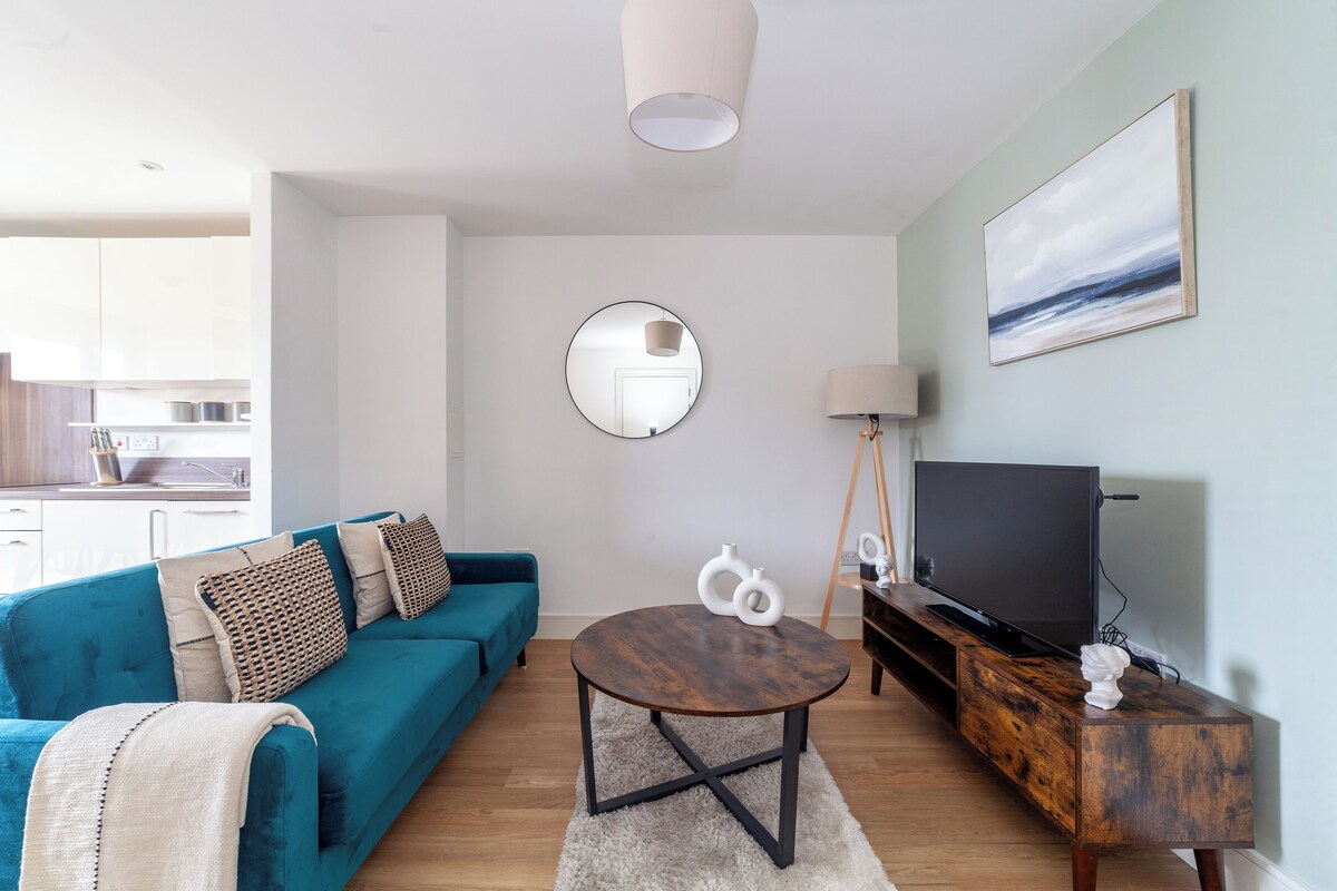 A modern living room with a teal sofa, a round coffee table, a TV on a wooden stand, and a kitchen in the background.