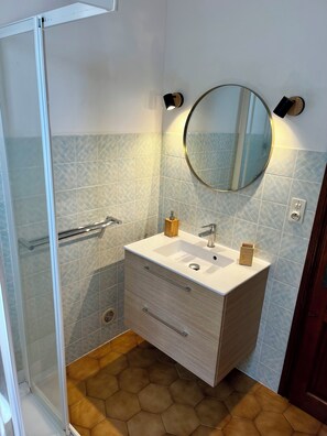A bathroom with a glass shower enclosure, a white sink with a round mirror above it, and hexagonal patterned tiles on the floor and walls.