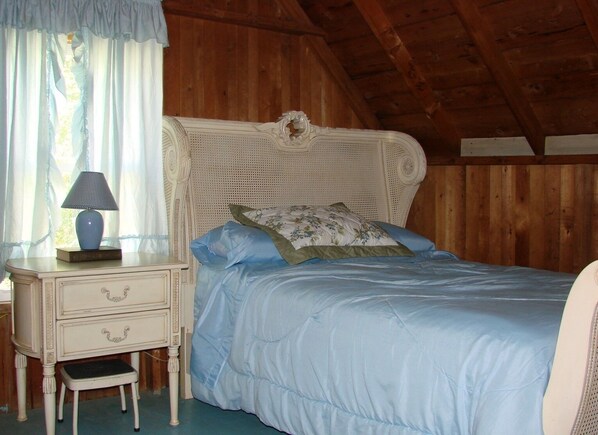 Un dormitorio con techo de madera, una cama tapizada en blanco con ropa de cama azul, una mesita de noche con lámpara y un taburete pequeño.