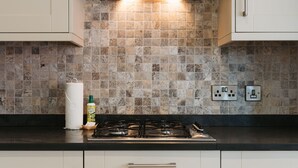 Eine Küche mit Gasherd, einer Edelstahl-Dunstabzugshaube und einer gefliesten Spritzschutzwand. Es sind Schränke mit Griffen und eine Arbeitsplatte mit einer Sprühreinigerflasche zu sehen.