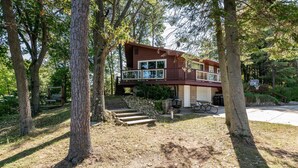 A two-story cabin with a balcony, surrounded by trees and a stone pathway leading to the entrance.