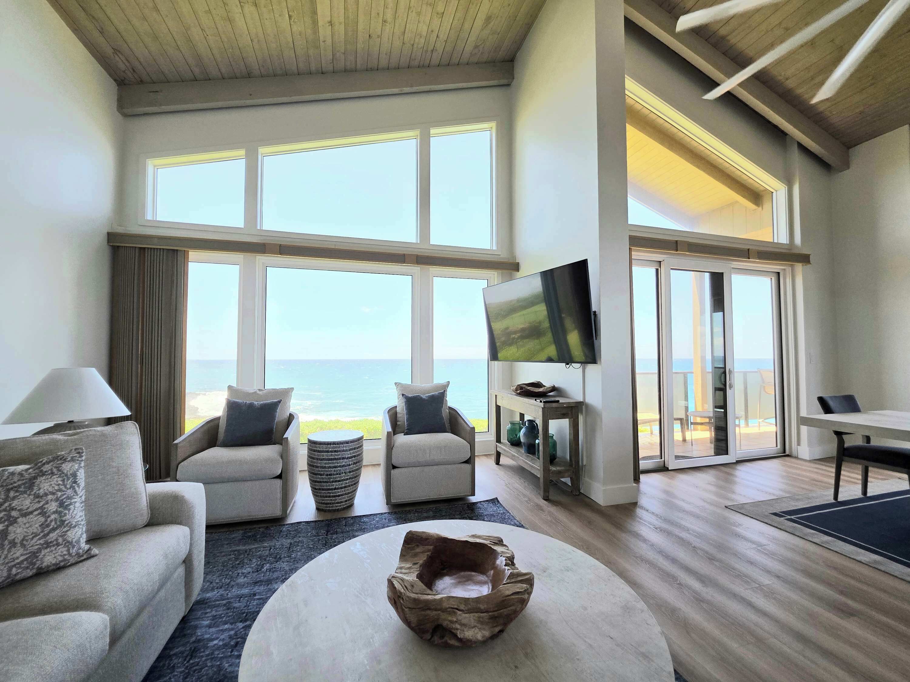 A spacious living room with a large window overlooking the ocean, a flat-screen TV mounted on the wall, and a cozy seating area with a round coffee table.