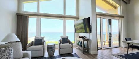 A spacious living room with a large window overlooking the ocean, a flat-screen TV mounted on the wall, and a cozy seating area with a round coffee table.