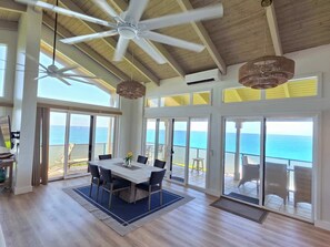 A spacious dining area with a large table and chairs, a ceiling fan, and a view of the ocean through large sliding doors.