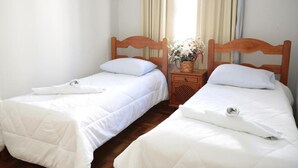 Two single beds with white bedding, a wooden headboard, a small bedside table with a vase of flowers, and a window with curtains. - HOTEL PLATINO (Sao Lourenco)