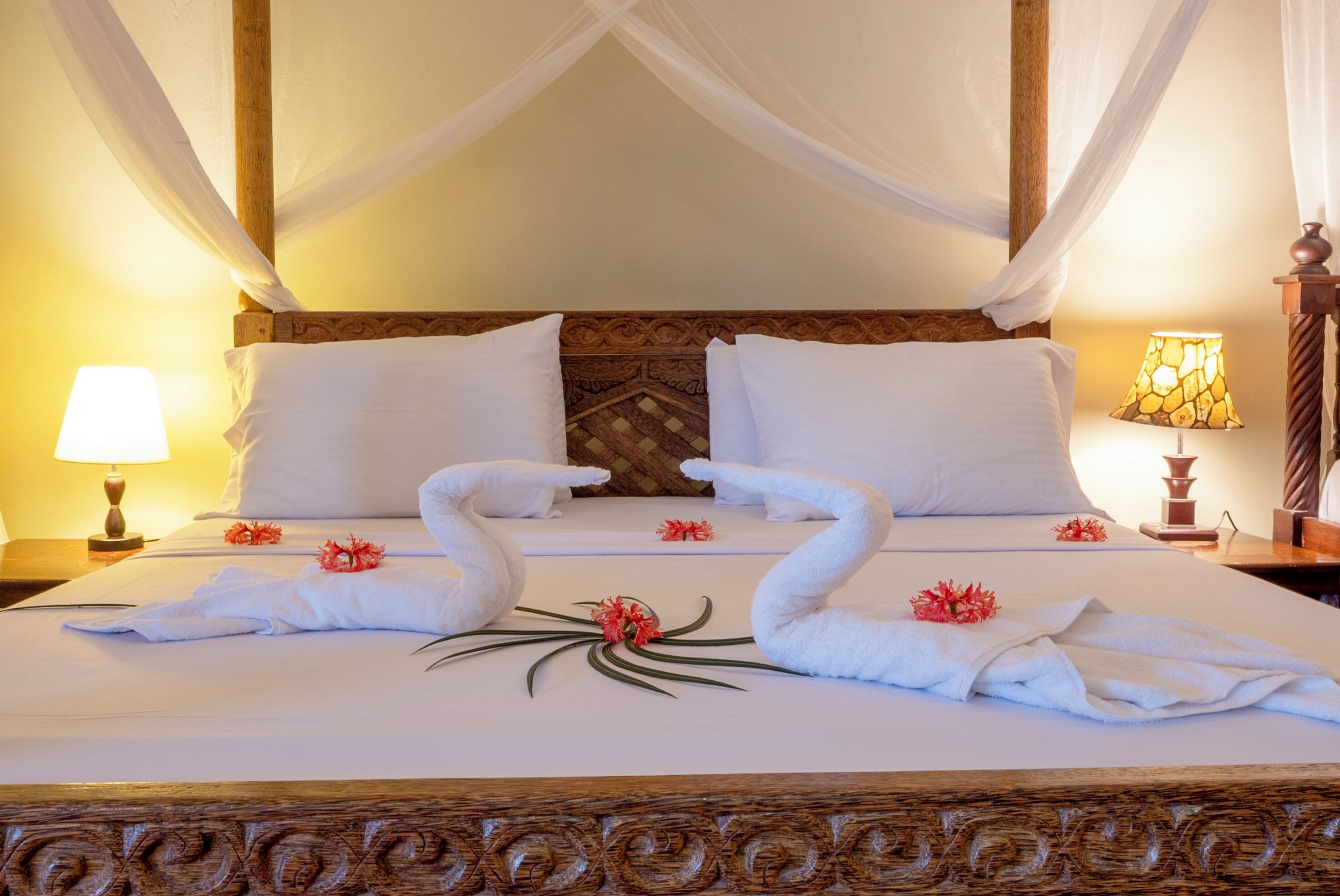 Uma cama com dossel, travesseiros brancos e toalhas dispostas em forma de cisne, decoradas com flores.