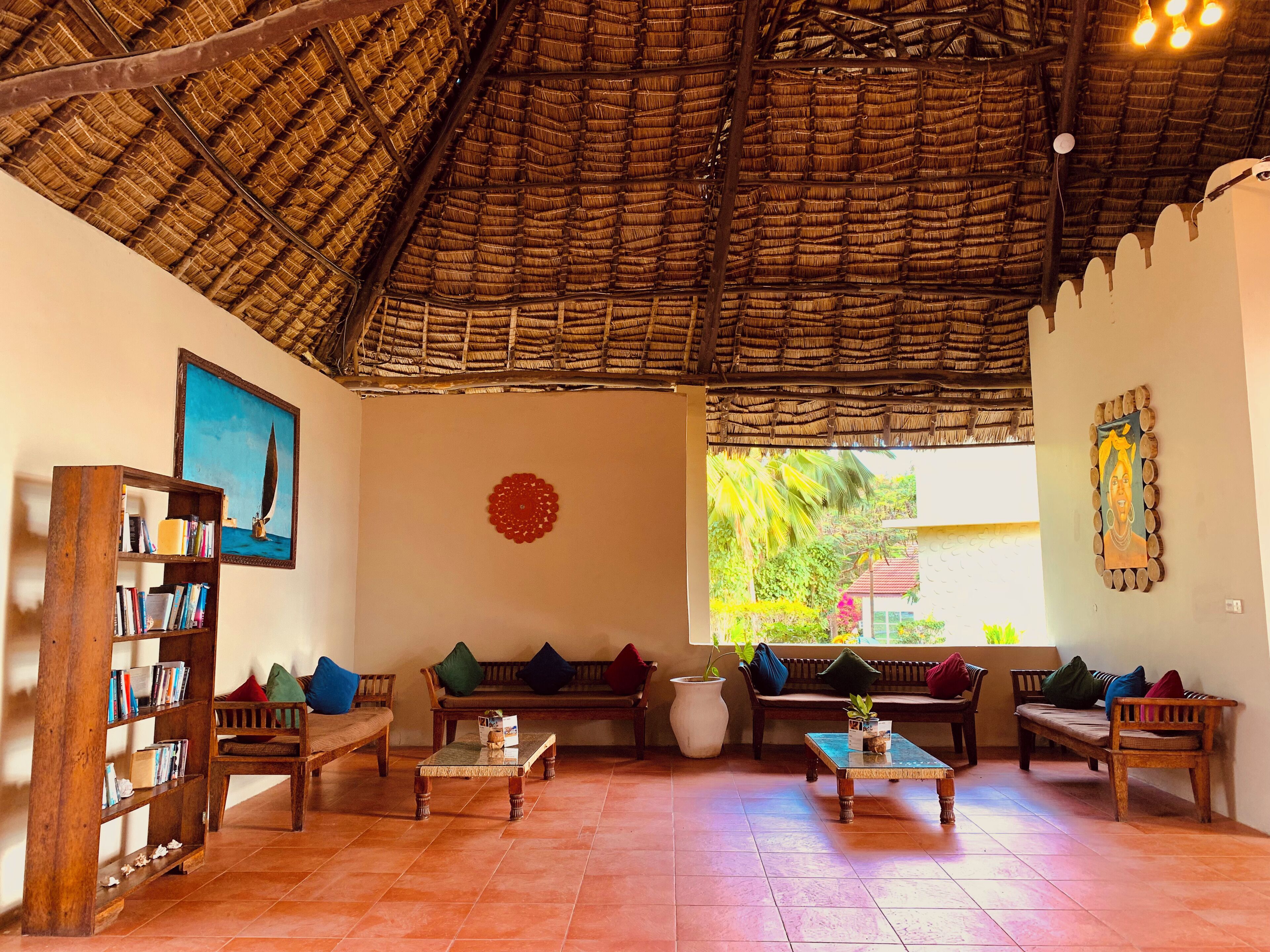 A room with a thatched roof, wooden furniture, a bookshelf, and a large window with a view of greenery.