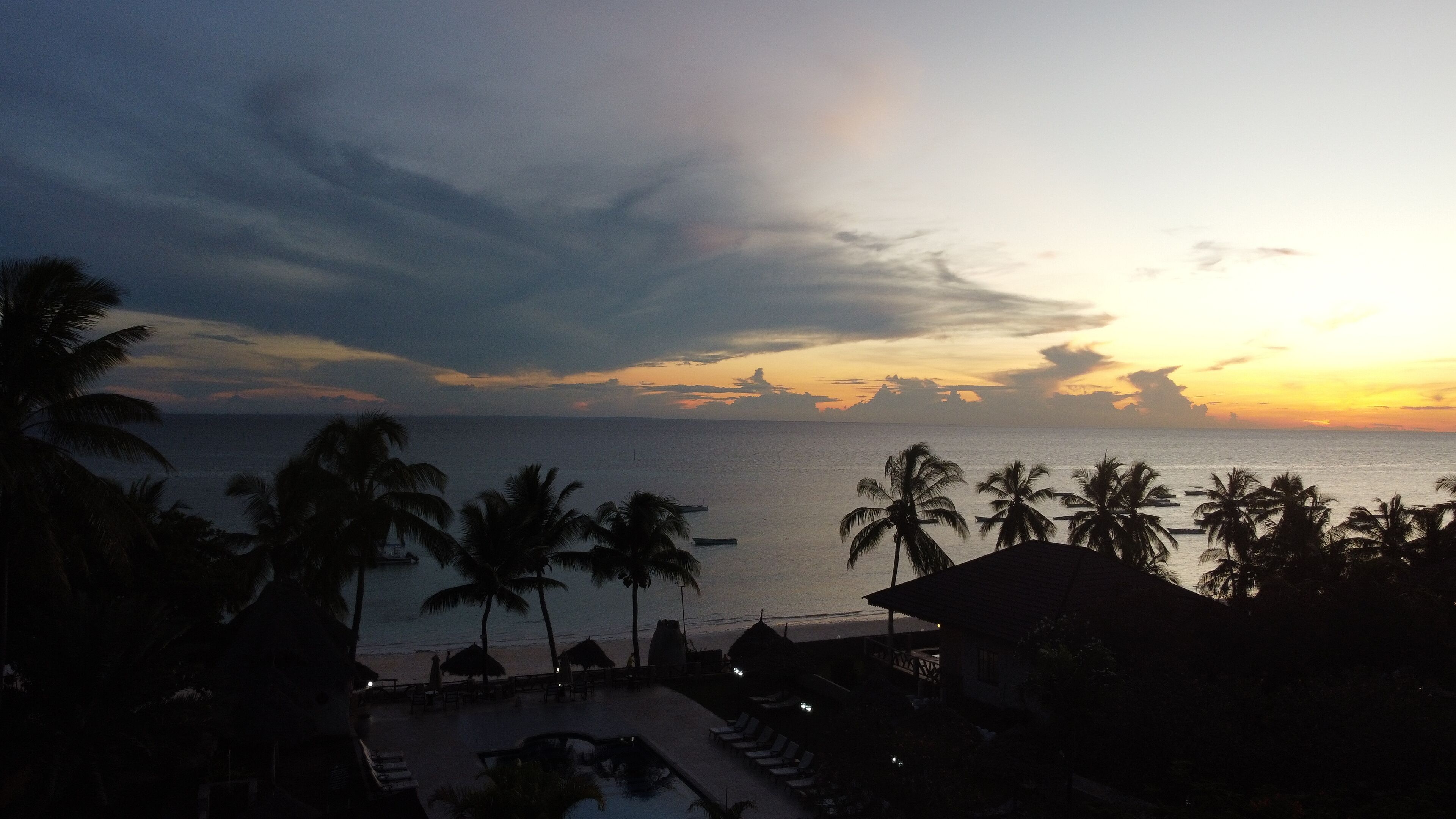 A resort with a pool, palm trees, and a view of the ocean at sunset.